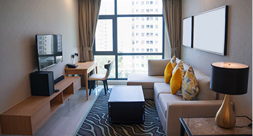 Sala de estar moderna e compacta com sofá cinza, almofadas amarelas, área de trabalho com mesa e cadeira de escritório, TV na parede e uma grande janela com vista para a cidade.