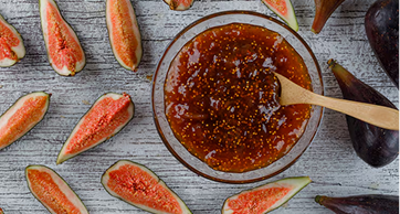 Vista superior de uma tigela de vidro com geleia de figo densa e brilhante, acompanhada por uma colher de madeira e fatias de figos frescos espalhadas sobre uma superfície rústica clara.