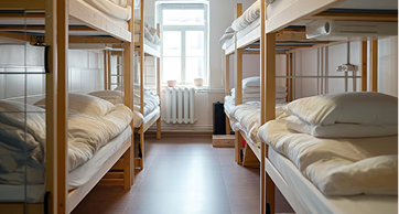 Interior de um dormitório compartilhado iluminado, com várias beliches de madeira arrumadas com lençóis e travesseiros brancos e uma janela ao fundo.