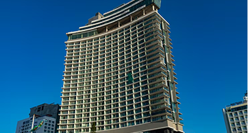 Fachada de um hotel moderno e alto com varandas de vidro, sob um céu azul limpo, representando opções de hospedagem urbana e corporativa.