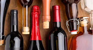 Close-up aéreo de diversas garrafas de bebidas dispostas verticalmente sobre uma superfície de madeira, incluindo vinhos, espumantes com lacre vermelho e garrafas com rolhas de cortiça e tampas de cerâmica.