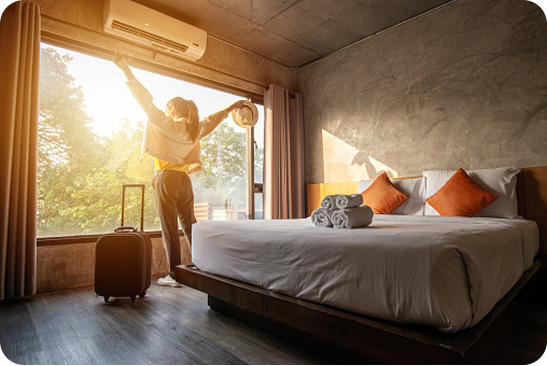 Mulher de costas abrindo as cortinas de um quarto de hotel moderno com paredes de cimento queimado, cama de casal arrumada e sol entrando pela janela, com uma mala de viagem ao lado.